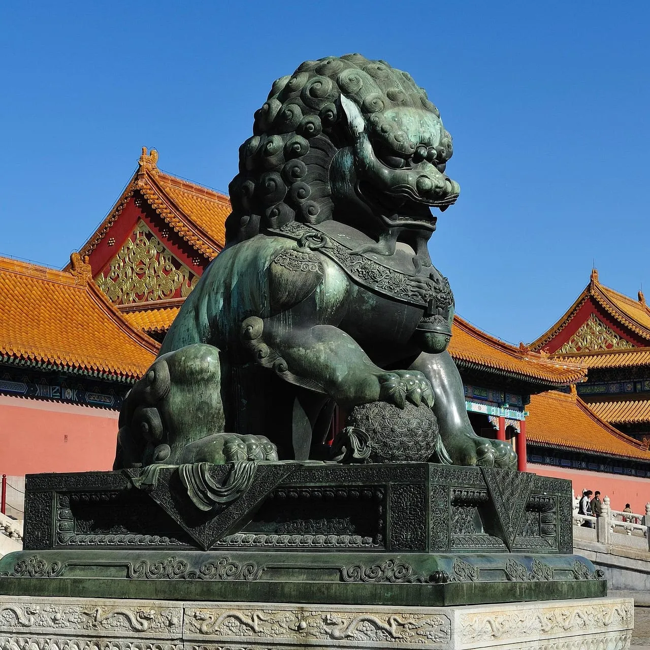 A bronze guardian lion in front of red walls and yellow roofs