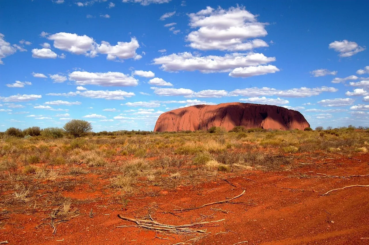 Uluru on the horizon across red earth and scattered bushes